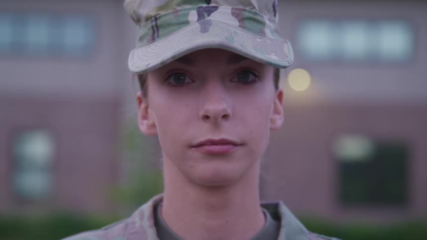 A woman in army uniform lifts her head and looks straight ahead while standing outside