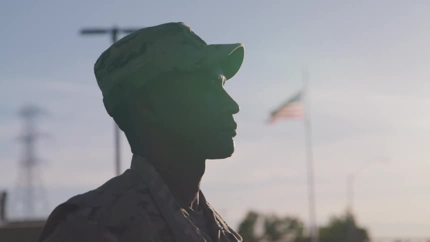 A military man stands in profile with the sun shining bright and an american flag flying on a pole