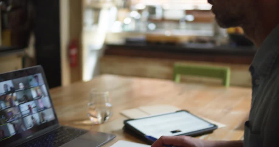 Man working from home talking on a video call throuhg a laptop while using a tablet and taking notes on a notepad