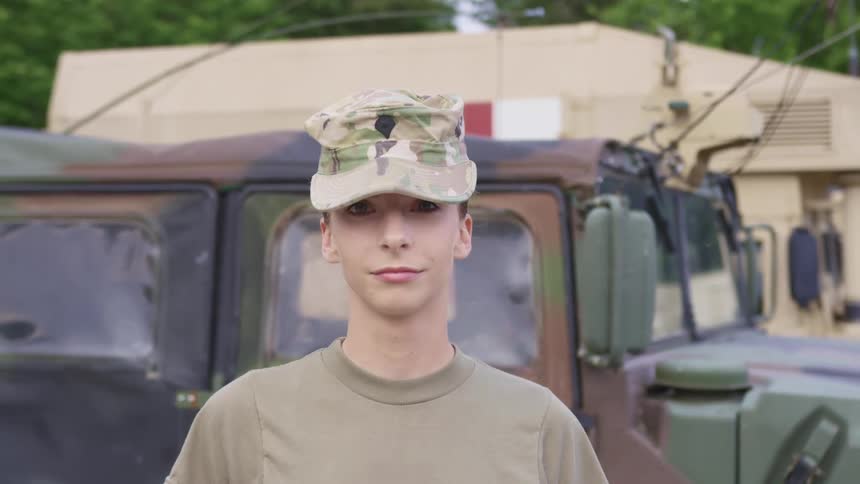 A young woman dressed in military wear stands in front of a military vehicle