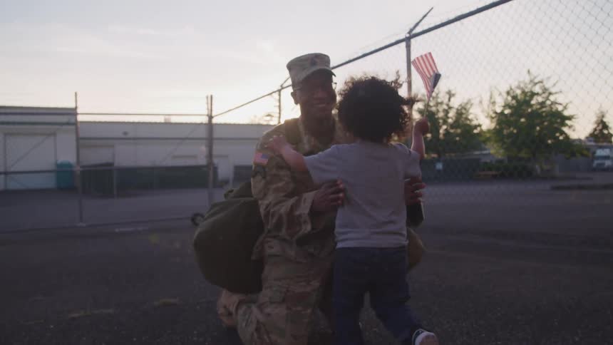 A toddler holds an american flag as he runs toward his soldier father in a parking lot and they hug