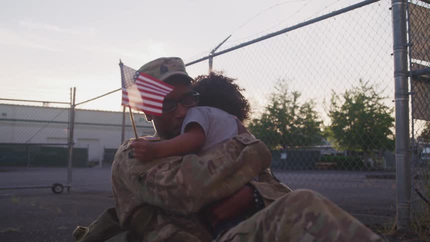 A soldier hugs his toddler who is holding a small flag
