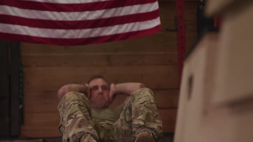 A male soldier doing situps in a camouflage uniform beneath an american flag