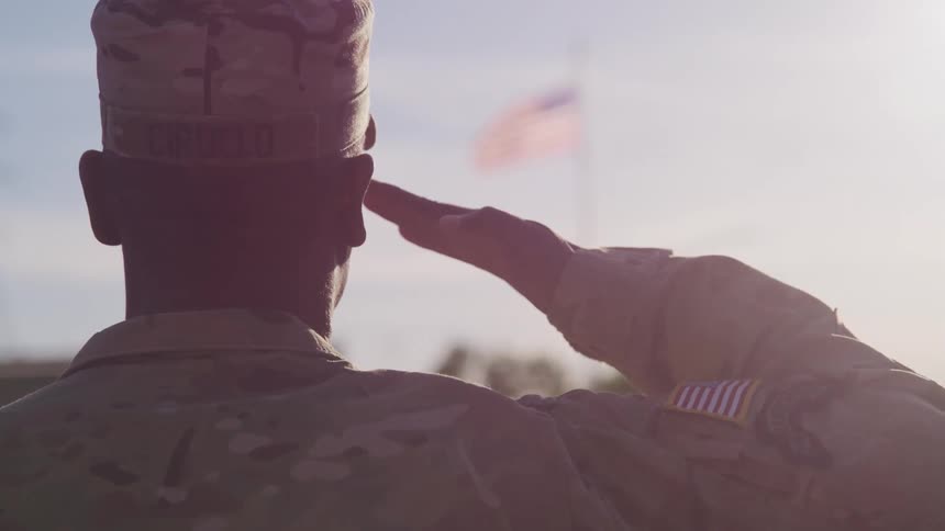 A soldier in uniform saluting an american flag outside on a sunny day
