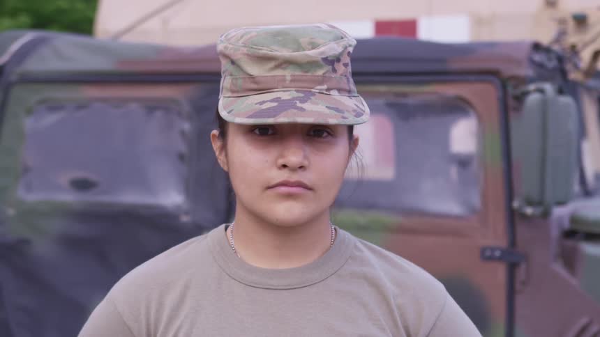 A woman with a military uniform standing and staring with a vehicle in the background