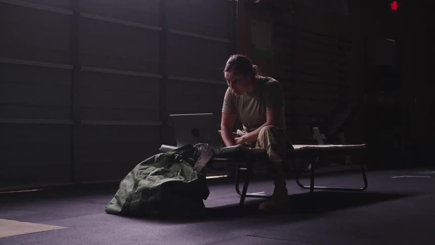 Young female army soldier sitting on a bed italking on a video call on a laptop