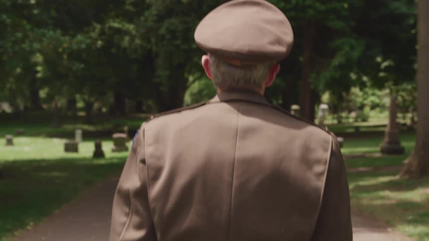 A gray haired man in a faded military uniform walks through a cemetery