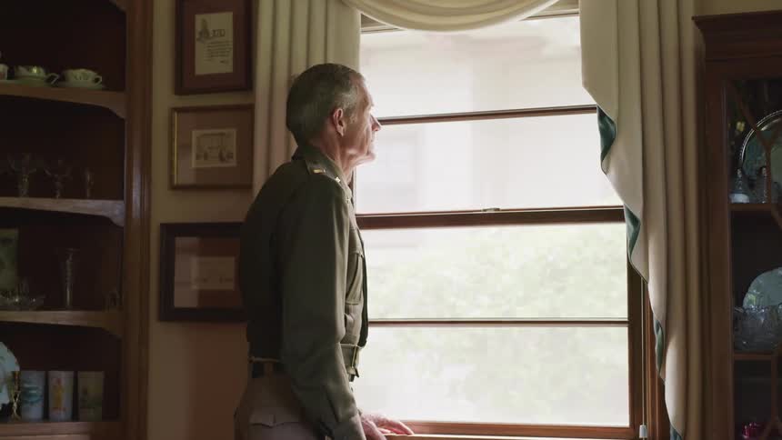 An elderly man stands and looks out of a window in his home