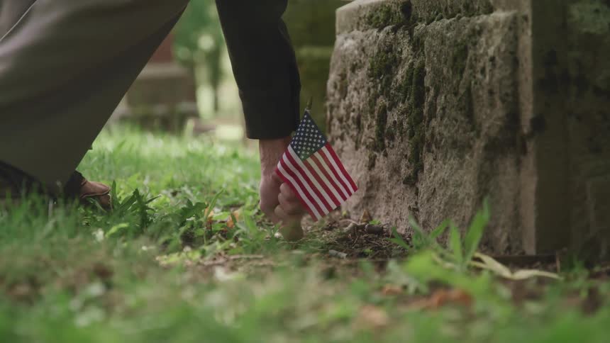 A soldier places an american flag in the ground in front of a cemetery headstone