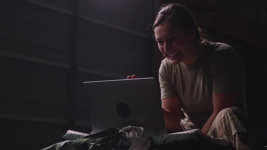 Woman wearing military uniform pants sits waving and smiling while video calling on a laptop