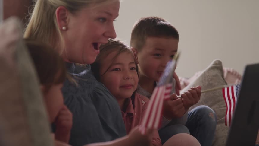 A mother sits in a couch with her children holding flags on her hands having a video call through a laptop
