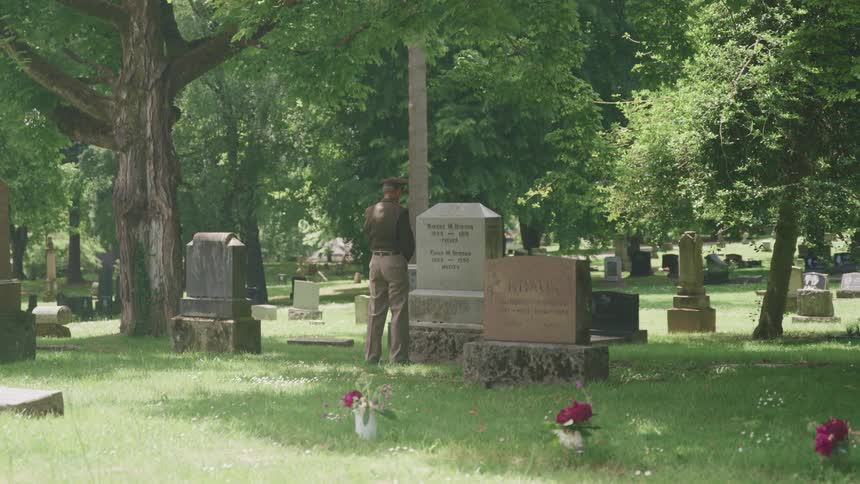 A man stands solemnly in front of a large tombstone in a cemetery during the day