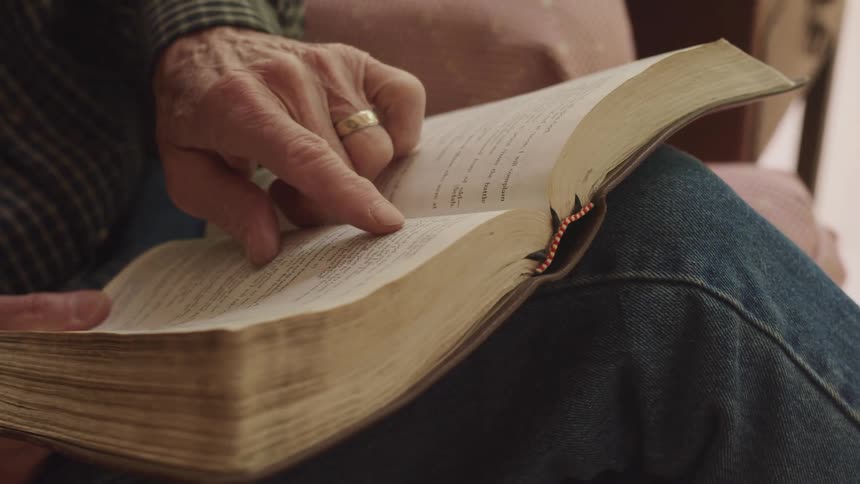 A man moves his finger across the page as he sits and reads a bible