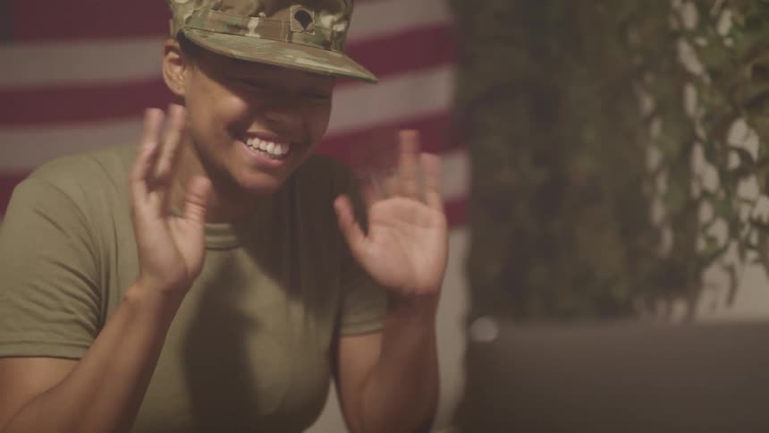 A woman with a military uniform in a room video chatting on a laptop with an american flag in the background