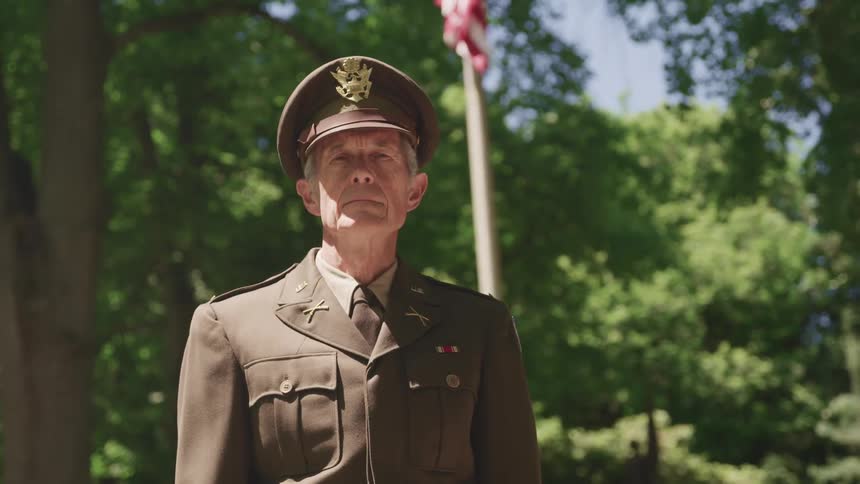Elderly man wearing an army uniform stands firm in front of an american flag outdoors looking at camera