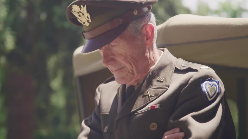 An elderly uniformed soldier stands outdoors with his head tilted in contemplation