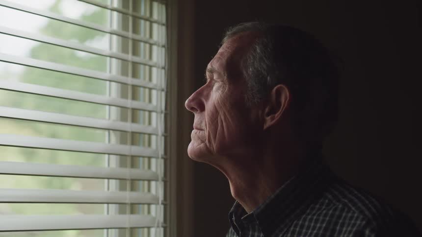 A man stands next to a window and looks through the open blinds