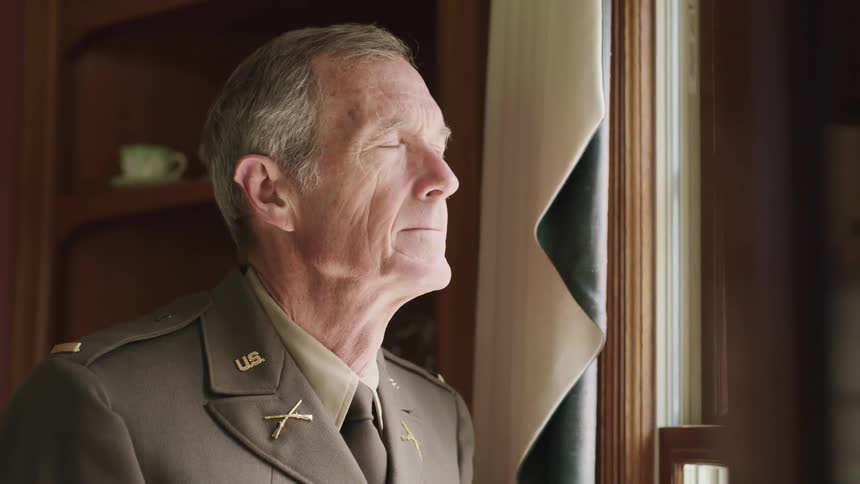 A man in a military uniform stands by his window and looks outside