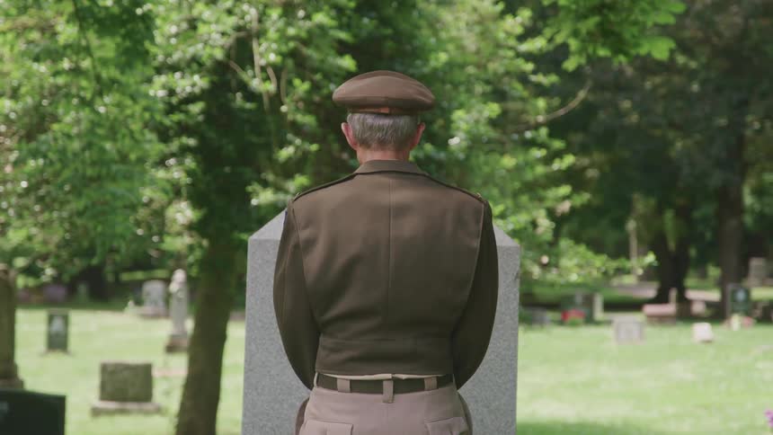 An older soldier in a military uniform standing in front of a grave at a cemetery