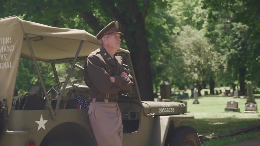A man in a military uniform leaning on a vehicle in a cemetery with trees in the background