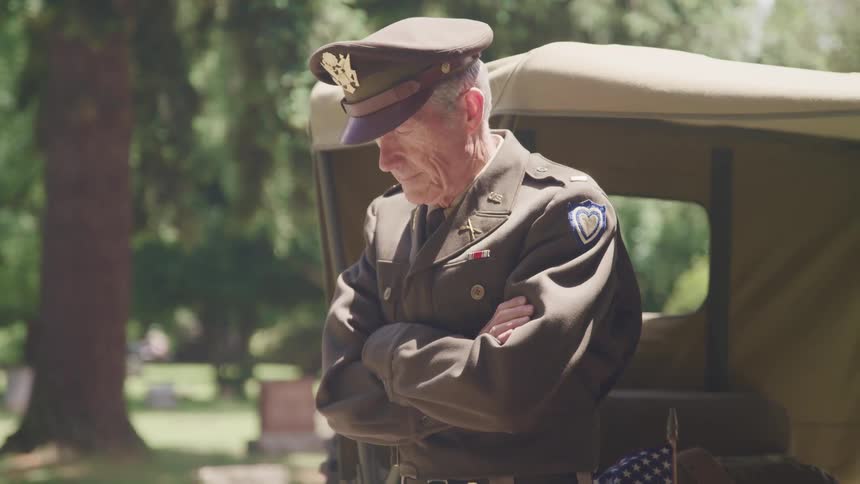 An elderly man in a military uniform stands with his headed bowed in a cemetary