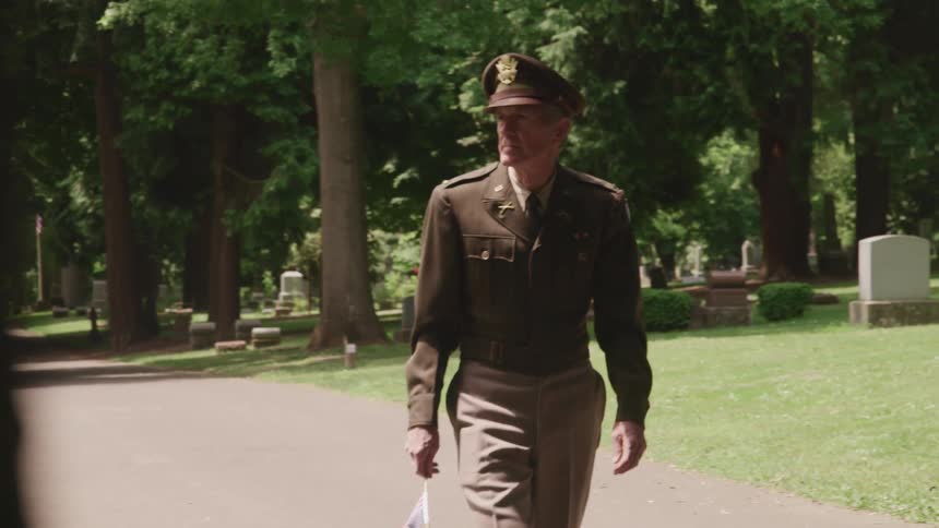 A man in full military uniform carries a small flag as he walks through a cemetery looking around as he goes
