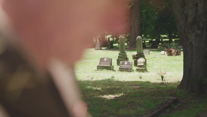 Person wearing uniform stands near shadow of tree looking down in cemetery