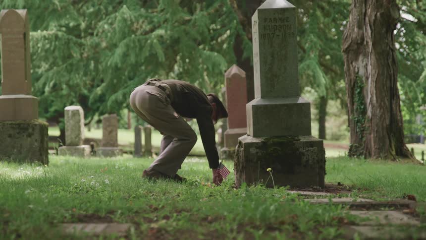 Elderly veteran army officer plants a small american flag near a tombstone in a cemetery