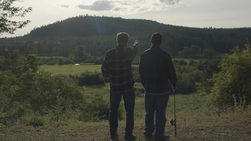 Father and son holding fishing rods stands on a hill looking at the mountains