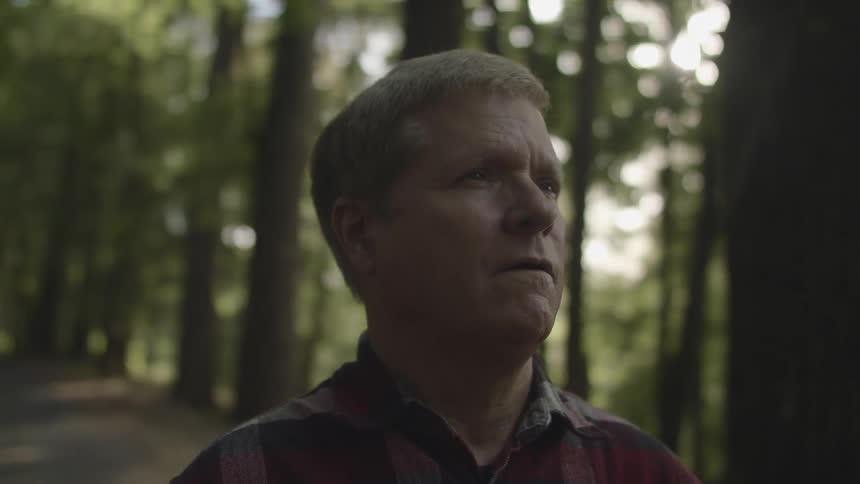 A man stands outside on a narrow pathway in a forest during the day staring ahead