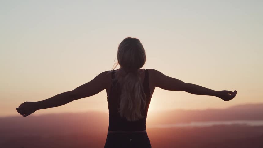 Woman faces sun sitting on horizon under red sky and stretches arms out
