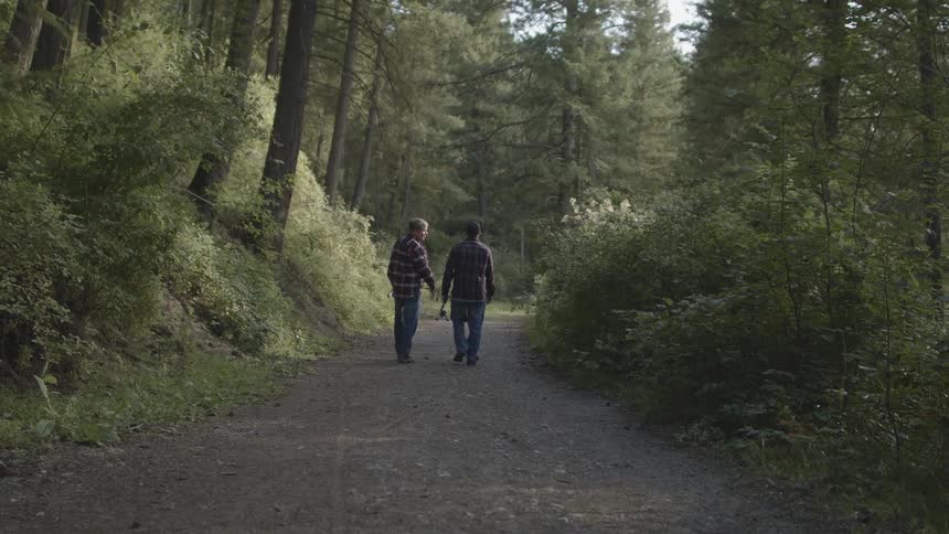 Two men with fishing rods walking in a pathway surrounded by trees with the sky and clouds in the background