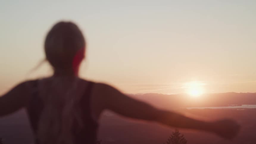 A woman standing outside with her hands stretched out with a lake a mountain range the sky and clouds in the background