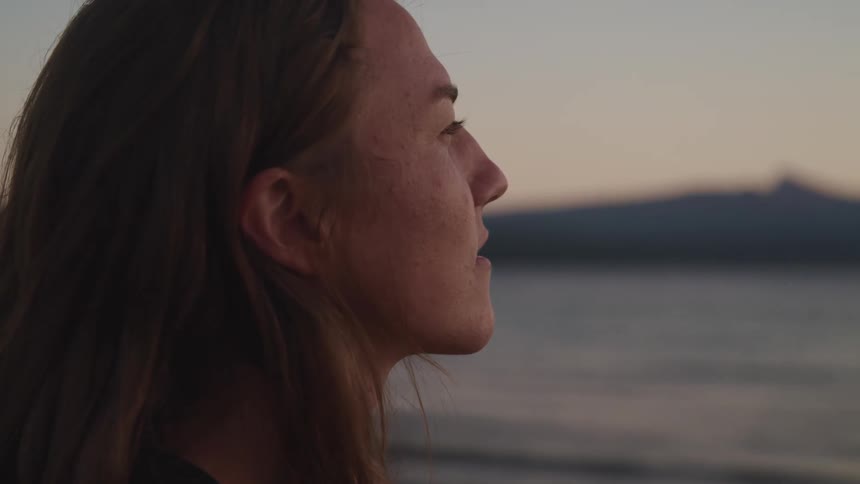 Young long haired woman standing on the coast of a beach looking at the sunset