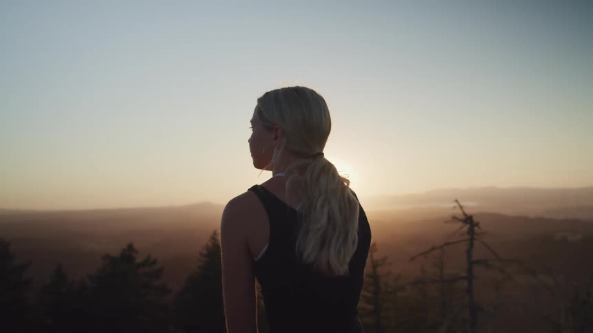 Blonde woman with ponytail and black top watches the horizon with sunset in the mountain