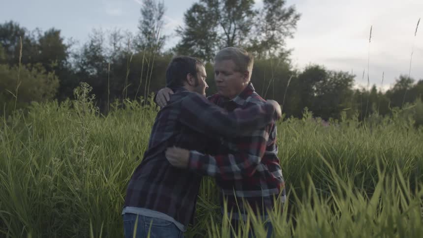 Two men standing in a grassy field hugging with trees the sky and clouds in the background
