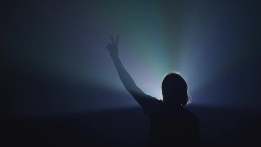 The silhouette of a person holding up their hand and giving the peace sign while a light shines