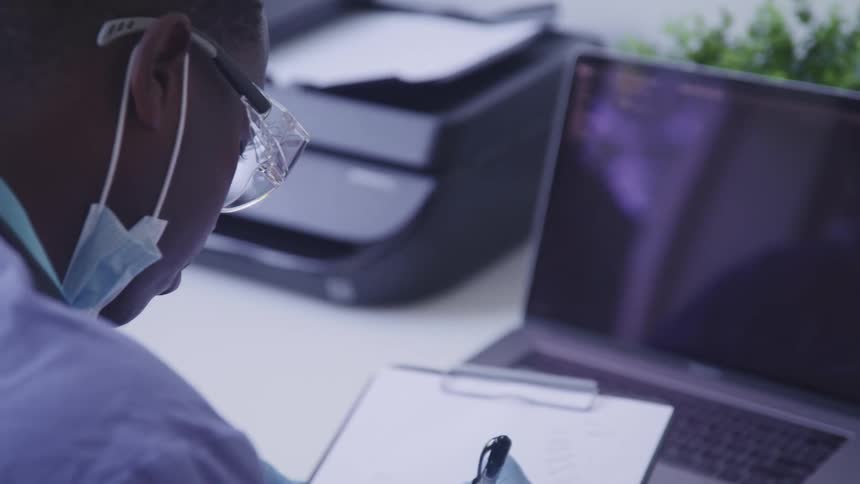 Medical person wearing mask makes notes near laptop