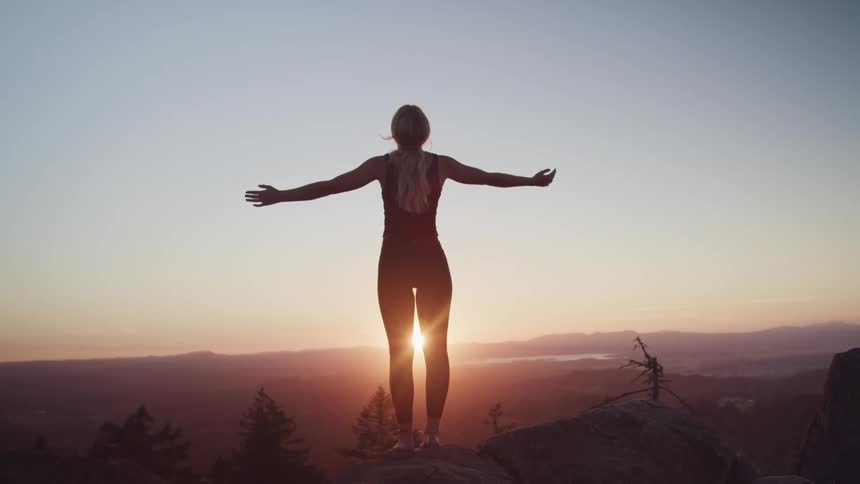 A woman stands at the edge of a canyon and stretches out her arms as the sun sets