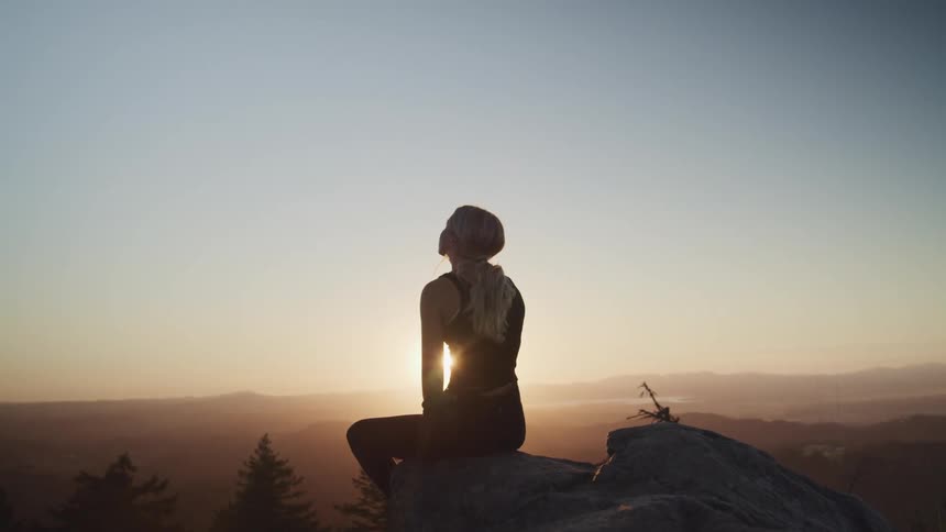 A woman sitting on a cliff with trees a mountain range the sky and clouds in the background
