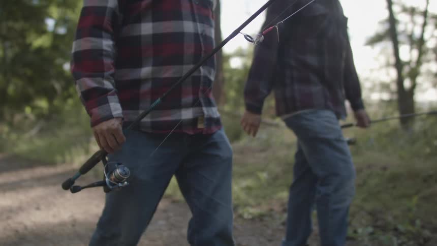 Two men carrying fishing rods walk down a path in a forest