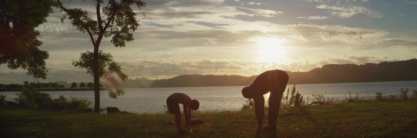 Father and son stretch on shore of lake as sun shines from over mountains