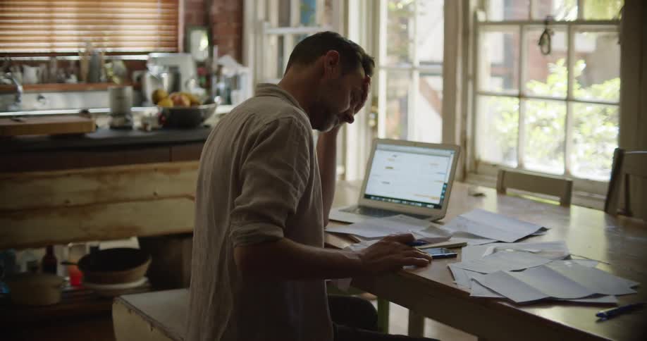 A man sits at a table with papers scattered around and a laptop open while he punches numbers into a calculator and looks out the window