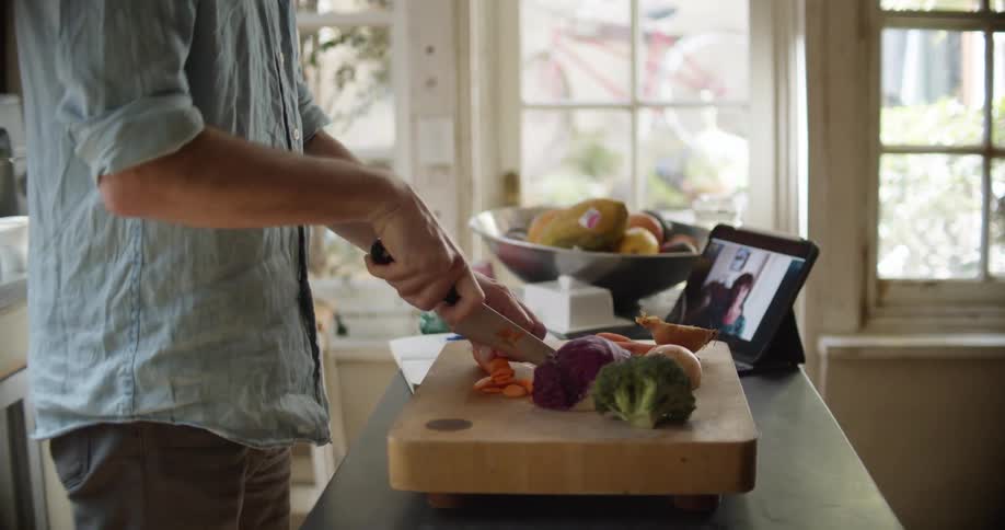 Man with blue shirt makes video call on tablet while cutting a carrot on cutting board in the kitchen