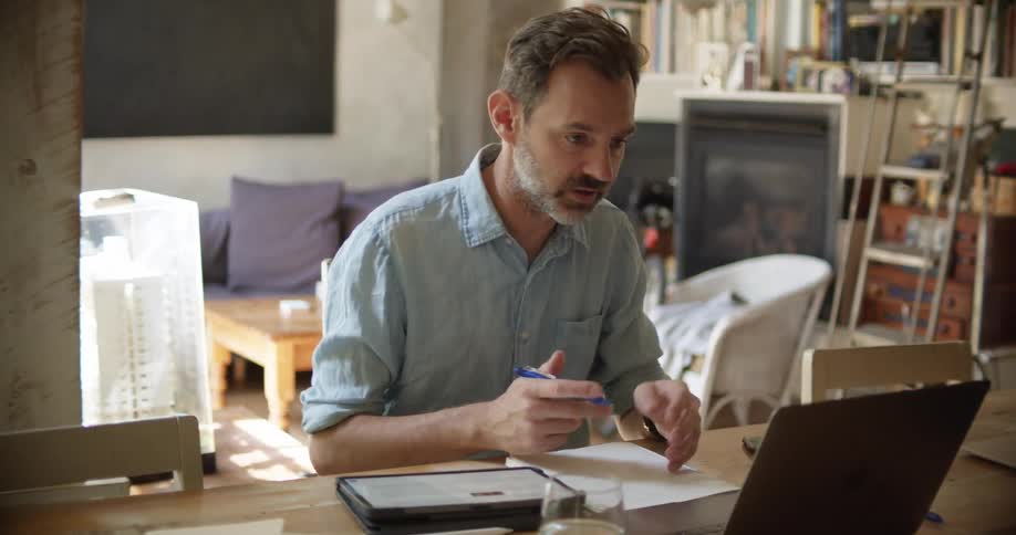 A man in a room sitting at a desk video chatting on a laptop writing on a paper with a pen