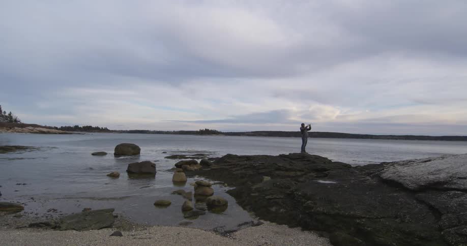 A man stands on a rock outcropping in a lake and takes a selfie