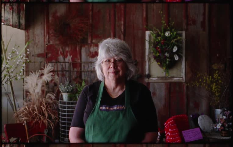 An elderly woman is standing in a room and wearing an apron
