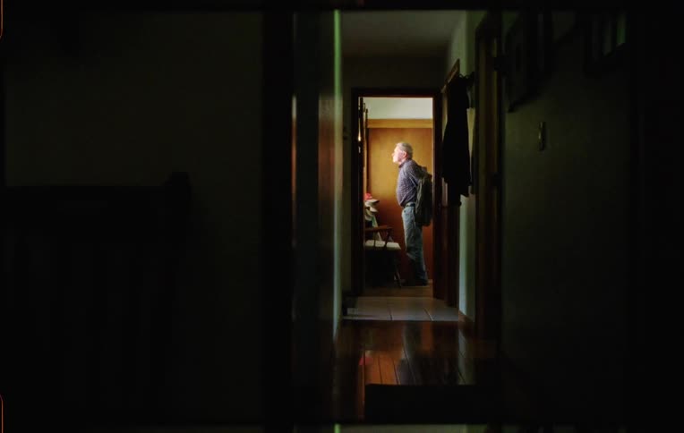 Elderly man walks into a room taking his hat and jacket off and sitting in a couch
