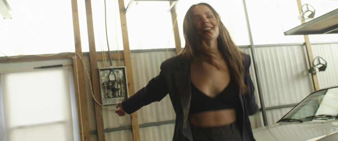 A young woman dances in front of a pair of cars in a large garage during the day