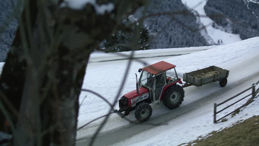 A tractor pulls a trailer along a snowy mountain road on a winter day